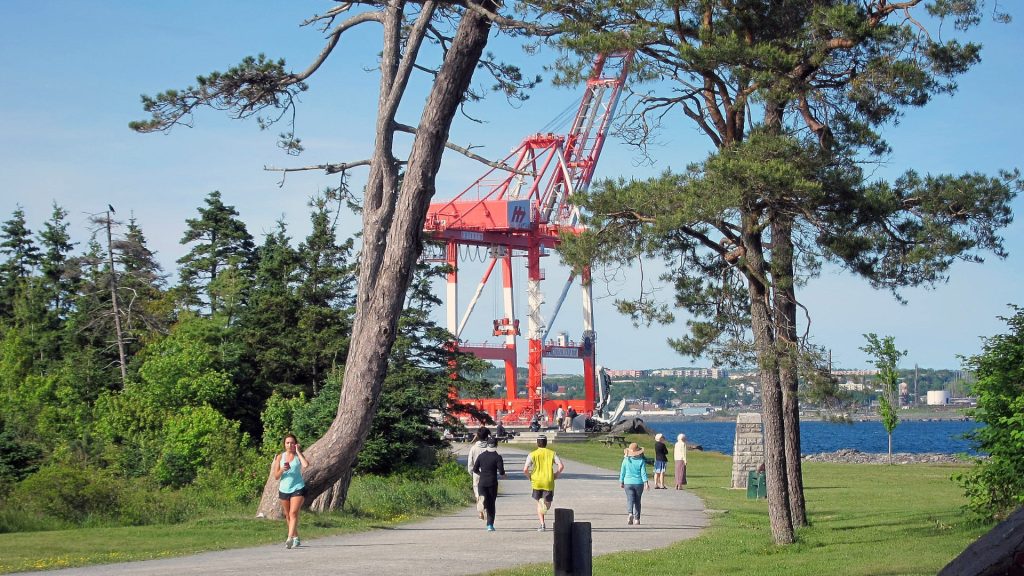 People walking and jogging along a trail in Point Pleasant Park with Halifax Harbour and port cranes in the background, Nova Scotia